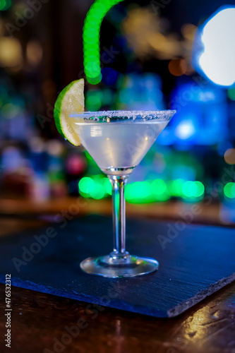In a funnel-shaped glass with a long stem, the alcoholic cocktail is opaque, opaque, with a slice of lime on the sugared edge of the glass. Filming of the bar menu against the background of bright