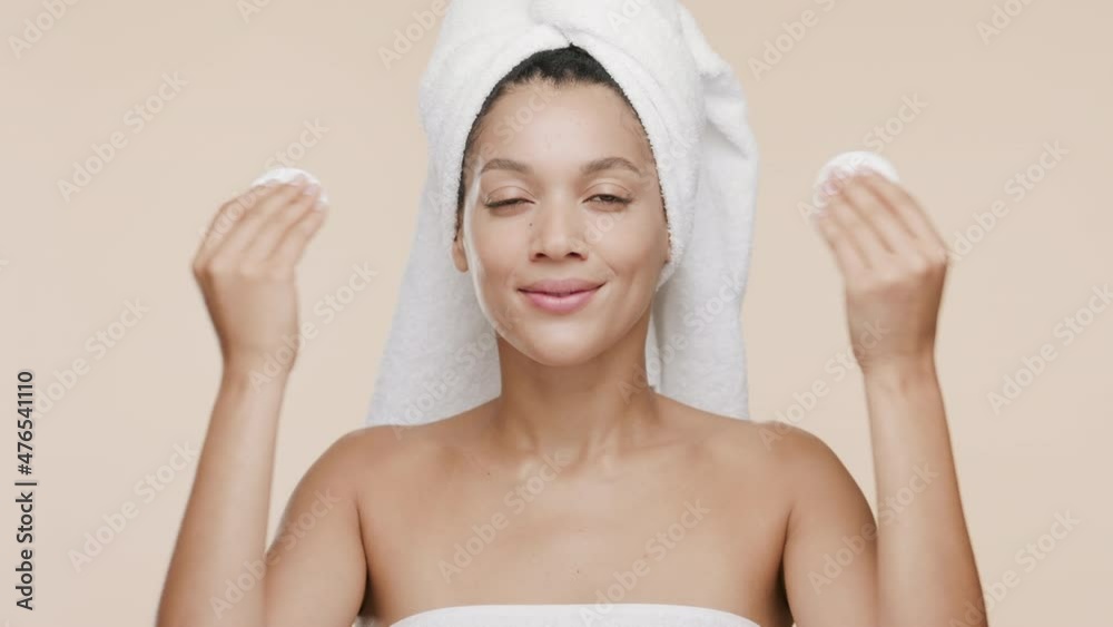 Beautiful young slim African American woman with a towel on hair and bare shoulders strokes nose and cheeks with cotton pads smiling wide for the camera on beige background | Acne prevention concept Beautiful young slim African American woman with a towel on hair and bare shoulders strokes nose and cheeks with cotton pads smiling wide for the camera on beige background | Acne prevention concept