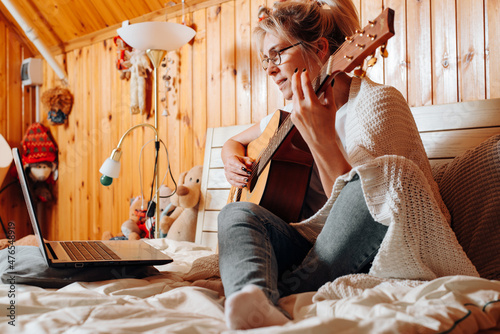 Learn music online, musical hobby and leisure concept. Middle aged caucasian woman playing acoustic guitar, studying music course, watching lesson on laptop while sitting on bed at home