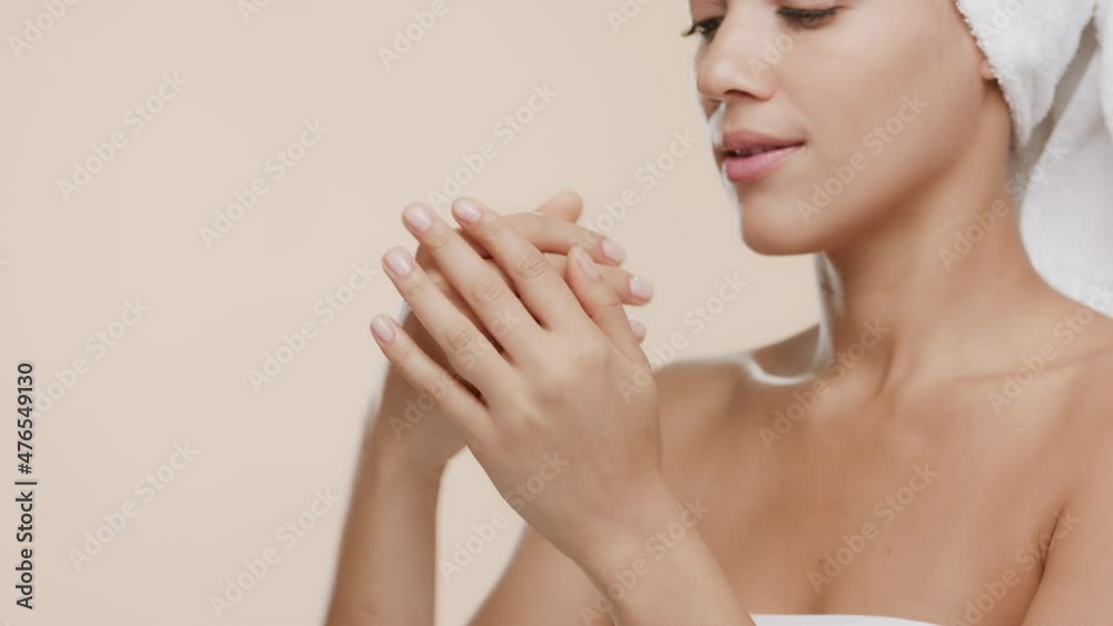 Cute slim African American young woman with a towel on her head and naked shoulders rubs her hands until dry and smells them enjoying the fragrance on beige background | Moisturizer commercial