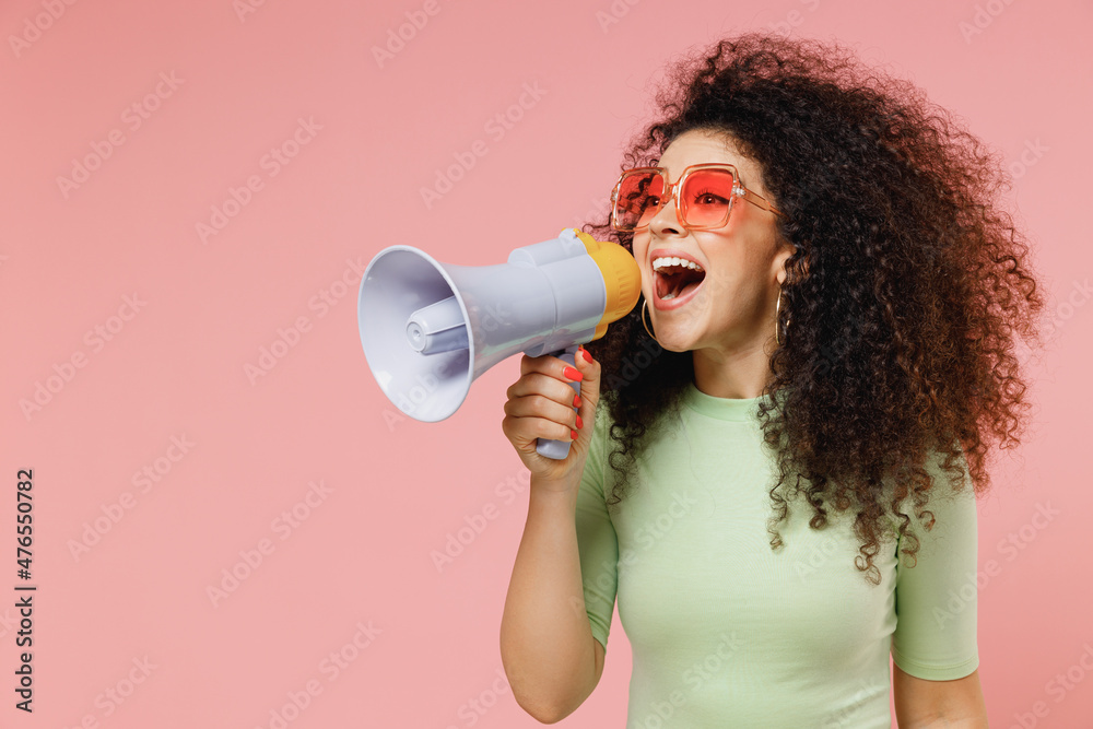 Exultant happy vivid young curly latin woman 20s wear mint t-shirt sunglasses hold scream in megaphone announces discounts sale Hurry up isolated on plain pastel light pink background studio portrait - obrazy, fototapety, plakaty 