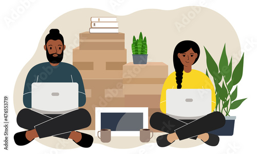 Couple sitting cross-legged with laptops, ordering delivery. Lots of things in cardboard boxes. The family is preparing to move to another house.