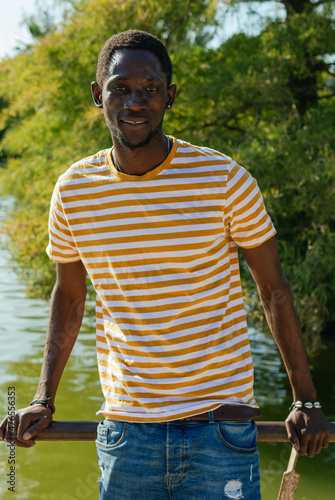 A portrait of an afro guy leaning on a railing wearing a plaid shirt with trees in the background.