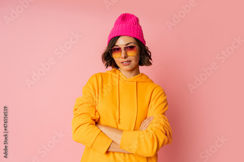 Woman in pink hat and orange hoodie posing on pink bacground. Stylish sunglasses.