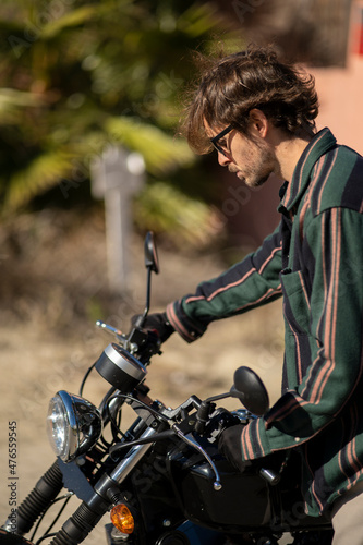 Adolescente hombre joven motero con camisa de cuadros elegante preparándose para viaje. Fondo de palmeras veraniegas. Chico elegante con gafas de sol y moto. Chico poniéndose casco de seguridad. photo