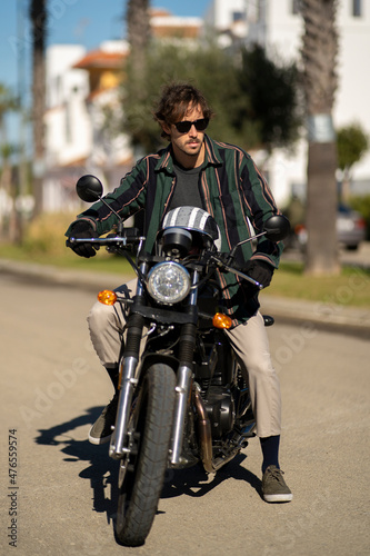 Adolescente hombre joven motero con camisa de cuadros elegante preparándose para viaje. Fondo de palmeras veraniegas. Chico elegante con gafas de sol y moto. Chico poniéndose casco de seguridad. © victor rangel