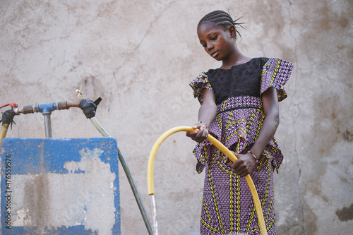 Canvas Print Pretty black African girl carefully filling a water tank with a yellow rubber ho