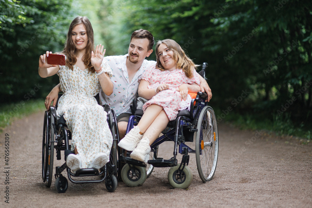 Positive women in wheelchairs and their male friend taking selfie on cell phone among green summer park. Concept of people with disability, gadgets and friendship. Positive women in wheelchairs and their male friend taking selfie on cell phone among green summer park. Concept of people with disability, gadgets and friendship.