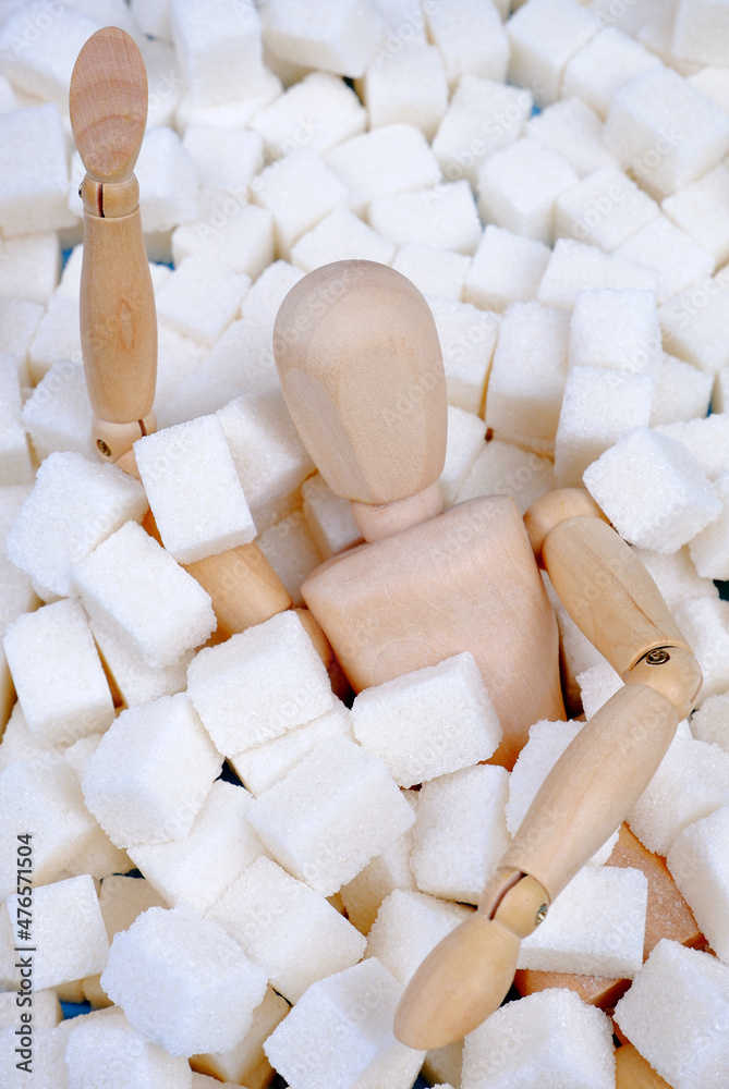 Fototapeta premium A wooden mannequin doll,strewn with sugar cubes,drowns in a pile of sugar.Unhealthy concept of sugar addiction,obesity and related diseases,diabetes,articles about the dangers of sugar.Vertical photo