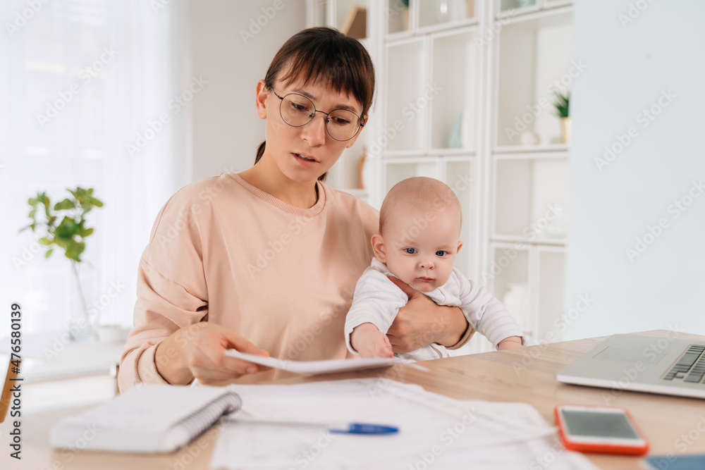 Beautiful serious business mom trying to work while spending time with her cute baby boy at home. Young working mother looking sadly at documents, sitting at her home office with her infant baby Beautiful serious business mom trying to work while spending time with her cute baby boy at home. Young working mother looking sadly at documents, sitting at her home office with her infant baby