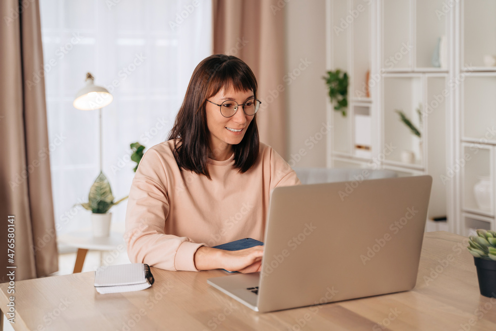 Pretty smiling woman in glasses working on laptop in home office, sitting at desk. Female freelancer working remotely. Student using computer for distance learning or retraining, typing on keyboard Pretty smiling woman in glasses working on laptop in home office, sitting at desk. Female freelancer working remotely. Student using computer for distance learning or retraining, typing on keyboard