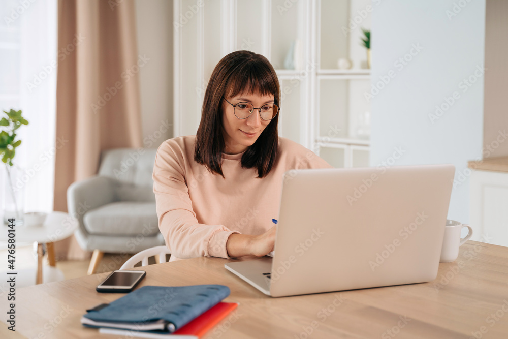 Smiling young woman wearing glasses working remotely, typing on laptop, looking at computer screen, sitting at desk in her home office. Female enjoying elearning process, studying online at home Smiling young woman wearing glasses working remotely, typing on laptop, looking at computer screen, sitting at desk in her home office. Female enjoying elearning process, studying online at home