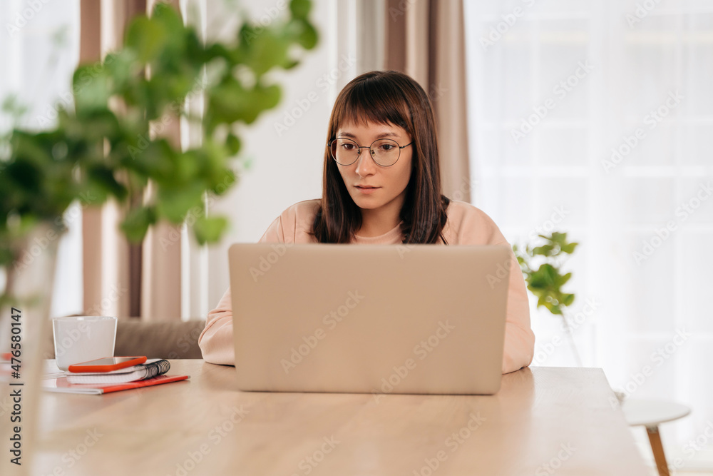Serious business woman wearing glasses working from home, looking at laptop screen, sitting at desk. Female doing remote distant job or studying get knowledge using online course. Self-education Serious business woman wearing glasses working from home, looking at laptop screen, sitting at desk. Female doing remote distant job or studying get knowledge using online course. Self-education