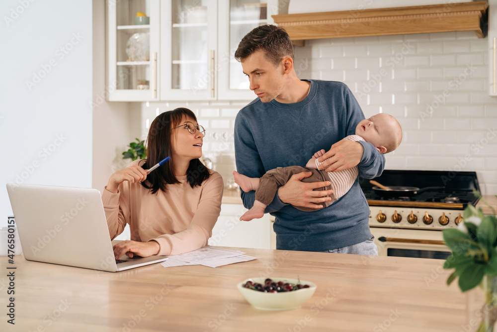 Young woman freelancer using laptop, working from home during maternity leave, family around. Young mother and father discussing family deals, making plans in kitchen while their cute baby is sleep Young woman freelancer using laptop, working from home during maternity leave, family around. Young mother and father discussing family deals, making plans in kitchen while their cute baby is sleep