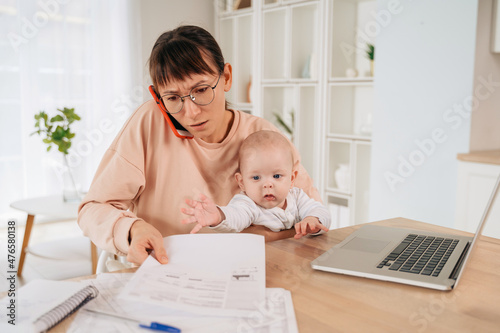 Busy single mother taking phone call and trying to solve family promlems while taking care of her little baby. Multitasking stressed mom talking on cellpphone, sitting at desk, holding her toddler photo