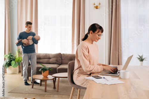 Concept of paternity leave instead of maternity one. Young woman working from home, using laptop in living room, while her husband taking care of their infant baby. Role of men in modern families 
