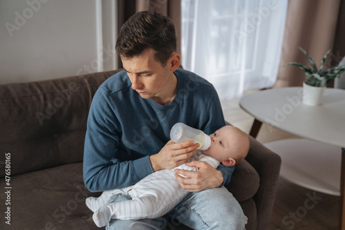 Unhappy young father feeding newborn baby with milk bottle on couch at home. Depressed single dad tired from sleepless night, fatherhood, take care of infant sun. Paternity and paternity leave concept photo