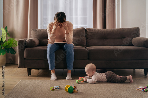 Young single frustrated woman hold her head with hands sitting on sofa in living room while her toddler son playing with toys on floor. Exhausted mom suffering from postpartum post-natal depression  photo