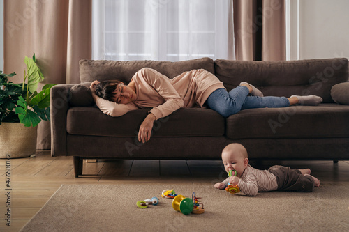 Fotografie, Obraz Young tired mother suffering from lack of sleep, sleeping on sofa while her little infant baby is playing on floor