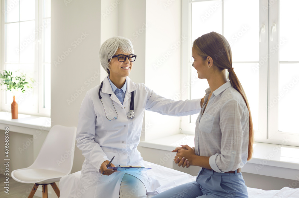 Fototapeta premium Smiling friendly female doctor touches patient's shoulder assuring that everything will be fine. Doctor sets young woman positive during conversations in hospital room. Concept of medical support.
