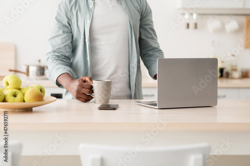 Morning routine concept. Unrecognizable african american man using laptop and drinking hot beverage in kitchen