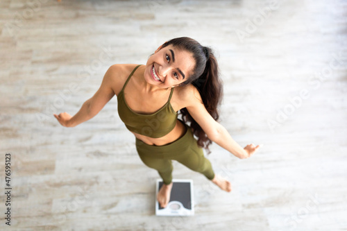 Above view of millennial Indian lady dancing on scales, choosing healthy lifestyle, happy with slimming result