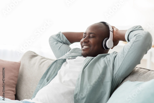 Happy black mature man in wireless headset reclining on sofa and smiling while listening to music with closed eyes