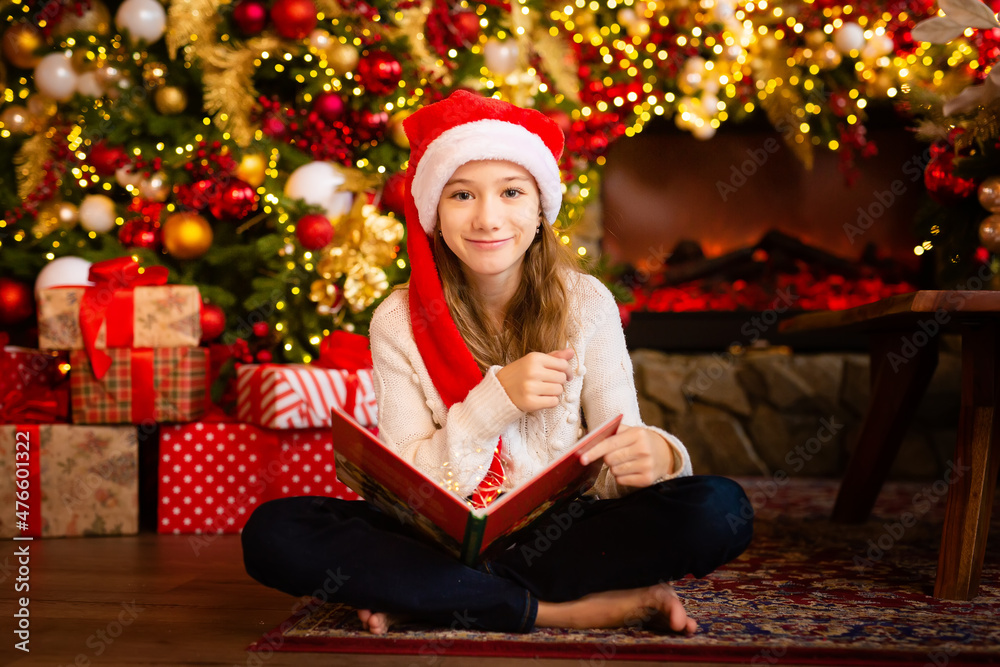 Little girl sitting near the Christmas tree, with a big book in her hands. Holiday decoration of the room, the Christmas tree