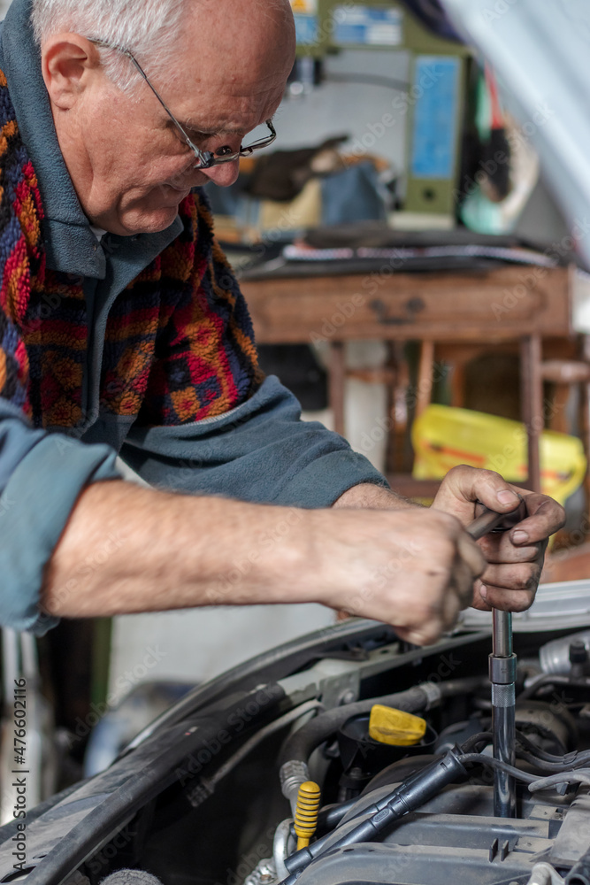 Portrait of a senior car mechanic changing spark plugs on his old car in the garage. Fixing spark plugs with a special long screwdriver. Old car mechanics and repair concept. Manual work. Portrait of a senior car mechanic changing spark plugs on his old car in the garage. Fixing spark plugs with a special long screwdriver. Old car mechanics and repair concept. Manual work.