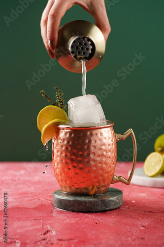 Pouring traditional american alcoholic beverage moscow mule in copper mug with lemon, thyme and huge ice cube from cocktail shaker on magenta and green background