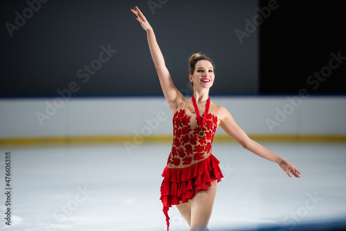 Photography positive figure skater in red dress and golden medal gesturing on ice arena