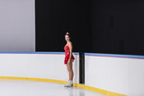 full length of young figure skater in red dress standing and looking at camera on ice rink