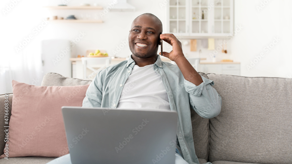 Mobile communication. Smaling african american man using laptop and talking on cellphone, sitting on couch Mobile communication. Smaling african american man using laptop and talking on cellphone, sitting on couch