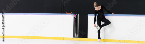 full length of figure skater in black bodysuit checking blade on ice skates near frozen ice arena, banner photo