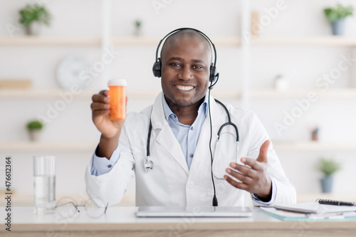 Doctor recommendation. Happy african american therapist in white coat showing jar of pills, having online consultation
