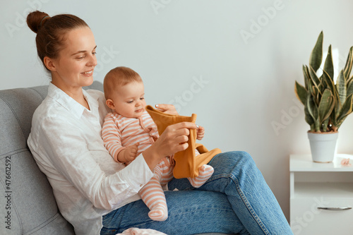 Fotografie Indoor shot of young adult Caucasian mother sitting on sofa with her infant daughter and holding child's clothing, having positive facial expression, admires new attire
