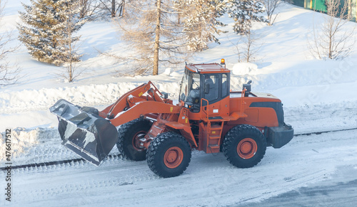 Big orange tractor cleans up snow from the road and loads it into the truck. Cleaning and cleaning of roads in the city from snow in winter