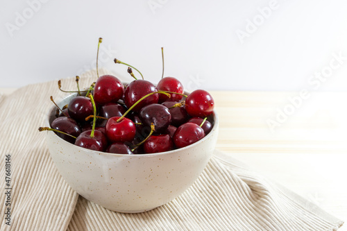 Organic red cherries in a ceramic bowl and a napkin on a light wooden table, healthy fruits as summer snack, copy space, selected focus