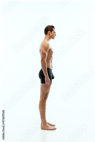 Profile view of young handsome shirtless sportive man wearing black boxer-briefs standing isolated on white studio background.