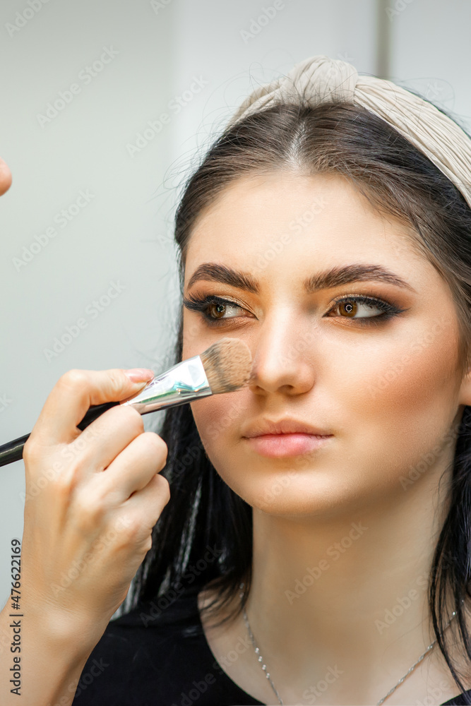 Fototapeta premium Closeup portrait of a woman applying dry cosmetic tonal foundation on the face using a makeup brush. Makeup detail
