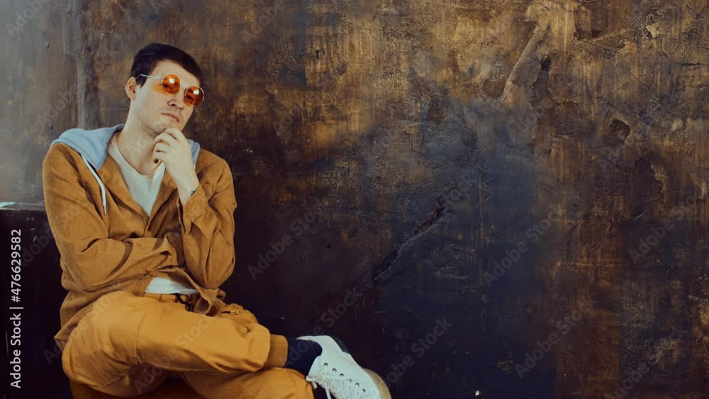Young handsome man in glasses sitting on chair and looking away. Stylish male on background of shabby brown wall.