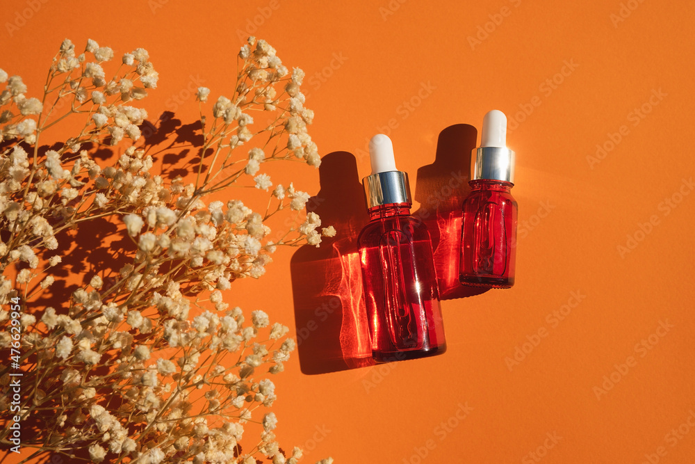 Red glass dropper bottle with silver lid on an orange background. Skincare products with beautiful daylight. Trending concept natural cosmetic with with dry plant. Beauty concept for face and body Red glass dropper bottle with silver lid on an orange background. Skincare products with beautiful daylight. Trending concept natural cosmetic with with dry plant. Beauty concept for face and body