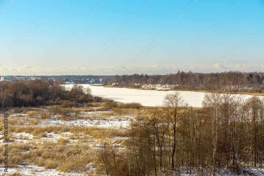 Panoramic winter landscape view with frozen river or lake covered with ice, reeds and some trees off the coast, small village and tower in background