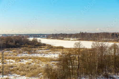Panoramic winter landscape view with frozen river or lake covered with ice  reeds and some trees off the coast  small village and tower in background