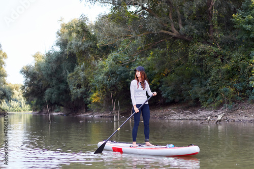 Caucasian young woman rowing on stand up paddle board, SUP alone in river near green trees