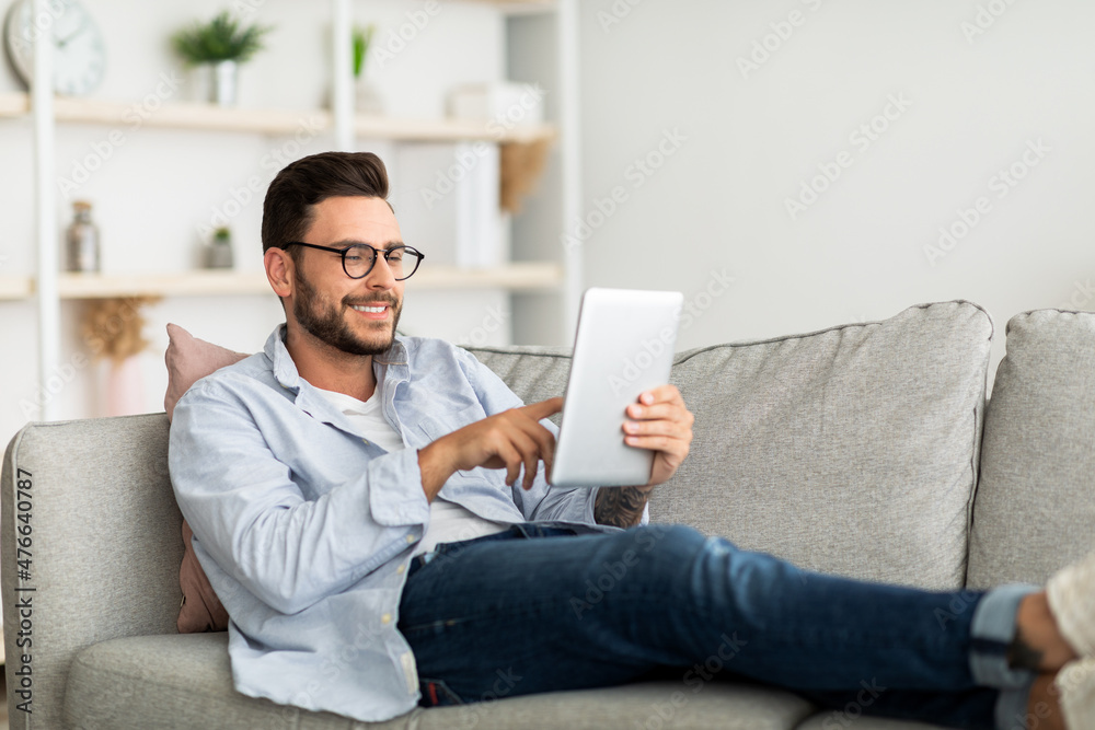 Technology concept. Smiling millennial man using digital tablet resting on couch at his modern apartment, free space