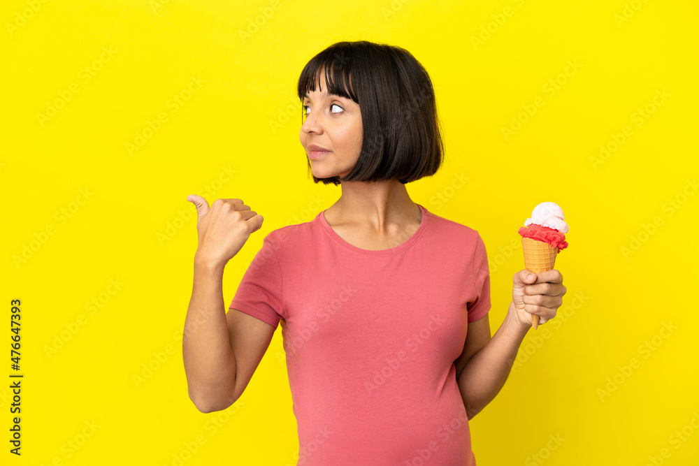 Pregnant woman holding a cornet ice cream isolated on yellow background pointing to the side to present a product Pregnant woman holding a cornet ice cream isolated on yellow background pointing to the side to present a product