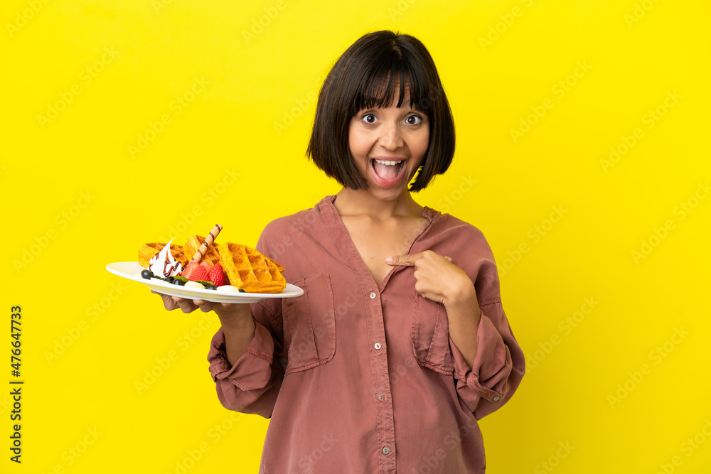 Pregnant woman holding waffles isolated on yellow background with surprise facial expression Pregnant woman holding waffles isolated on yellow background with surprise facial expression