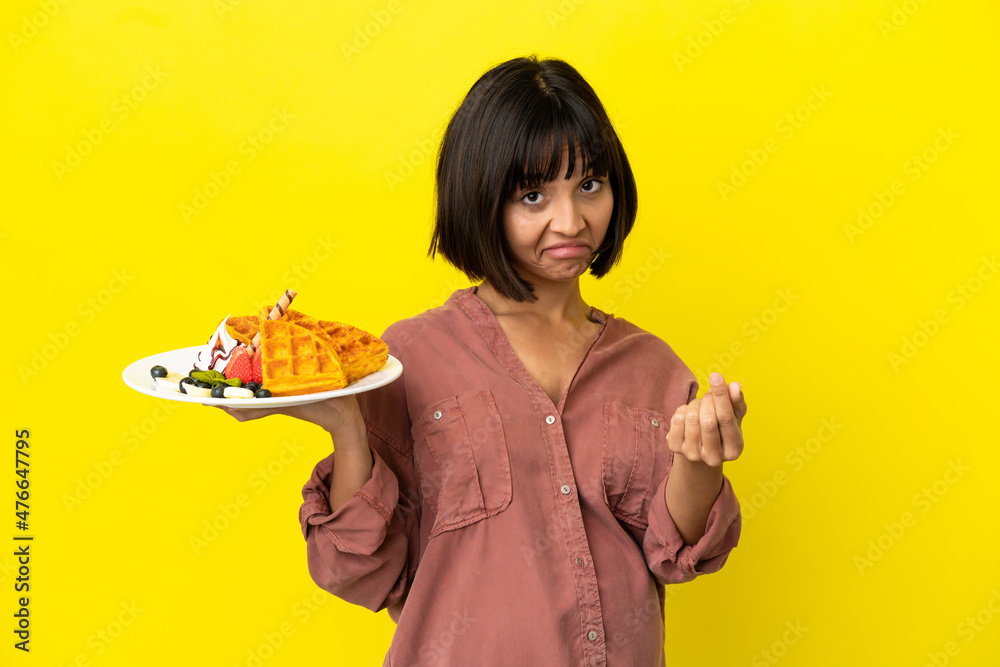 Pregnant woman holding waffles isolated on yellow background making money gesture Pregnant woman holding waffles isolated on yellow background making money gesture