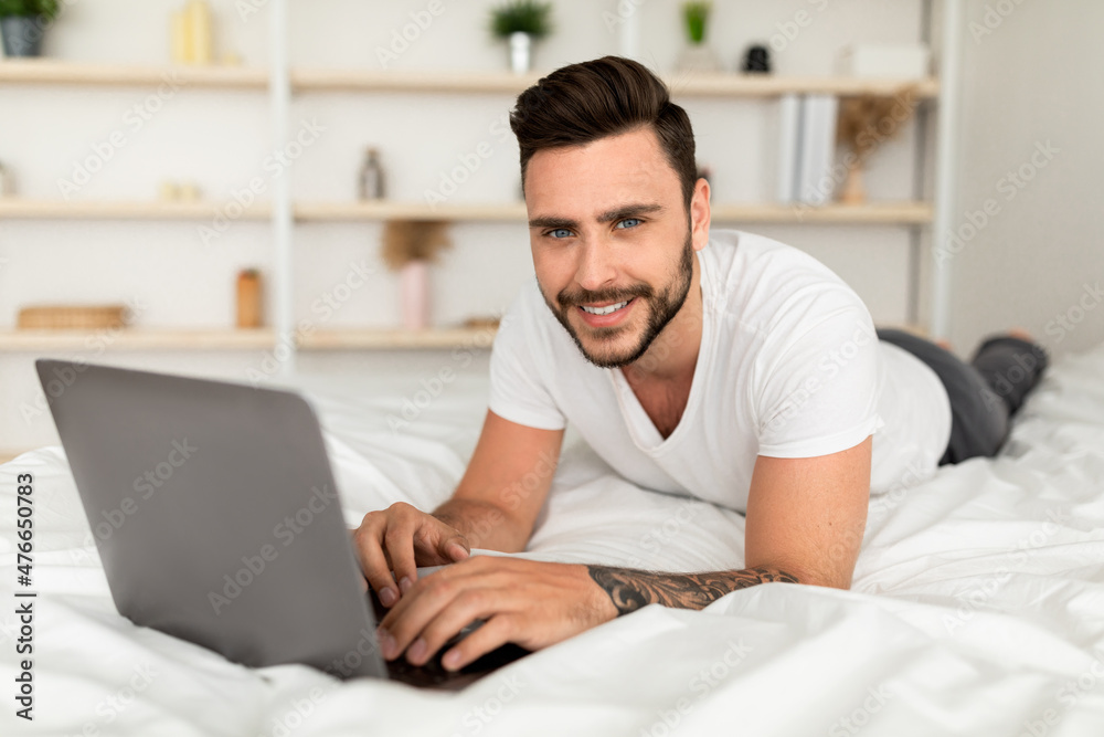 Job in bedroom, browsing on internet. Smiling millennial man woke up, working on laptop, lying on white bed in bedroom Job in bedroom, browsing on internet. Smiling millennial man woke up, working on laptop, lying on white bed in bedroom